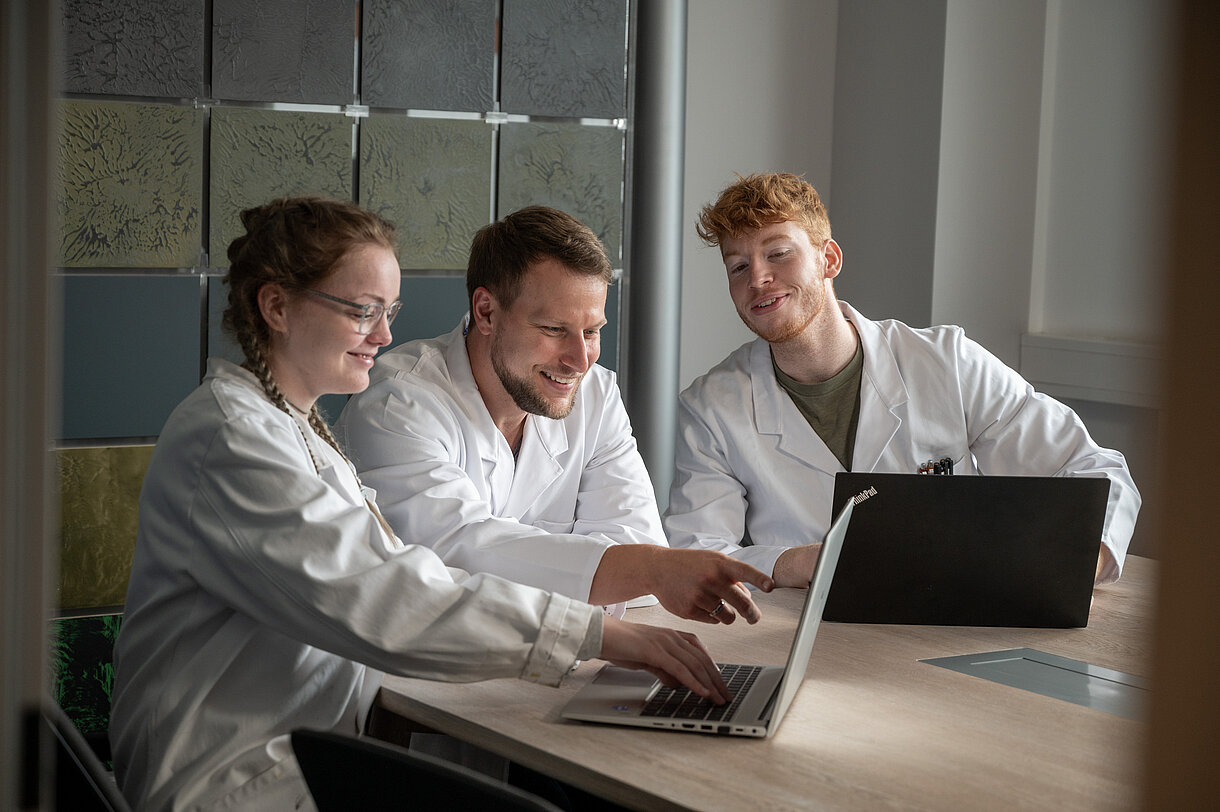 Drei Personen in Arbeitskleidung vom Labor besprechen etwas vor dem Laptop